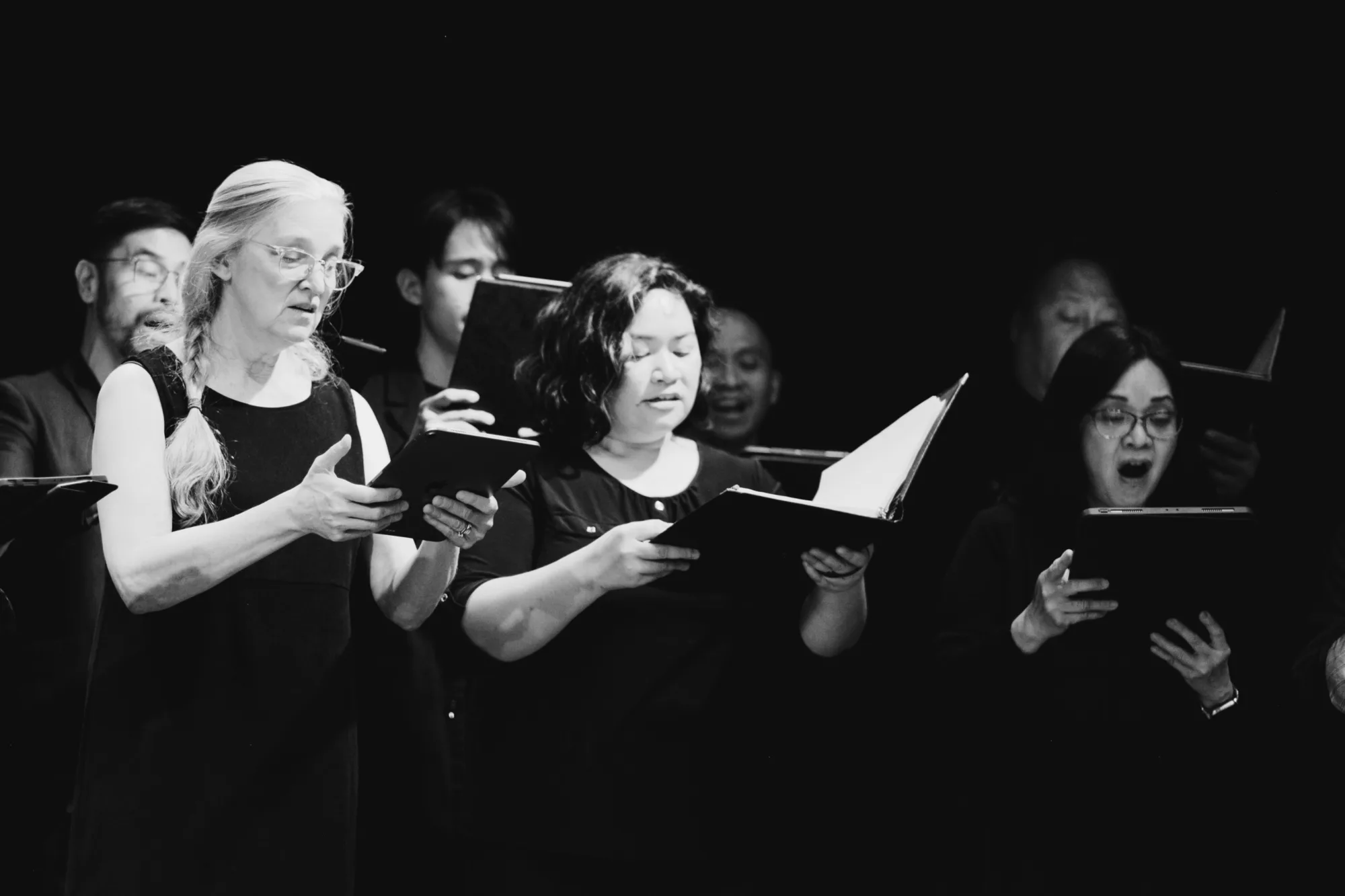 Close-up of choir singers mid-song holding black music folders against a dark stage
