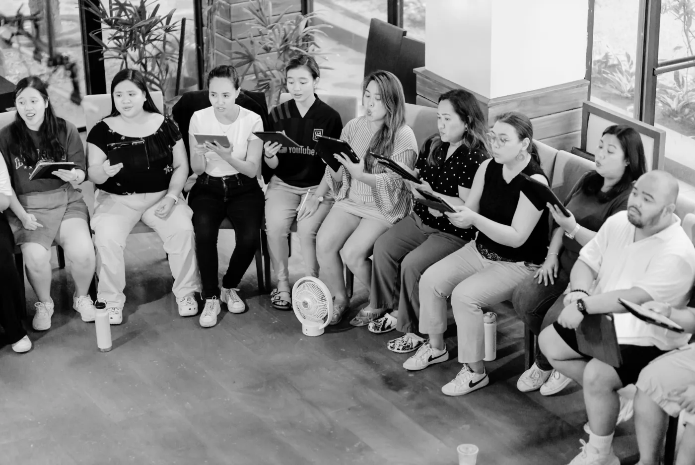 A choir seated in rows in a bright rehearsal room, holding music folders