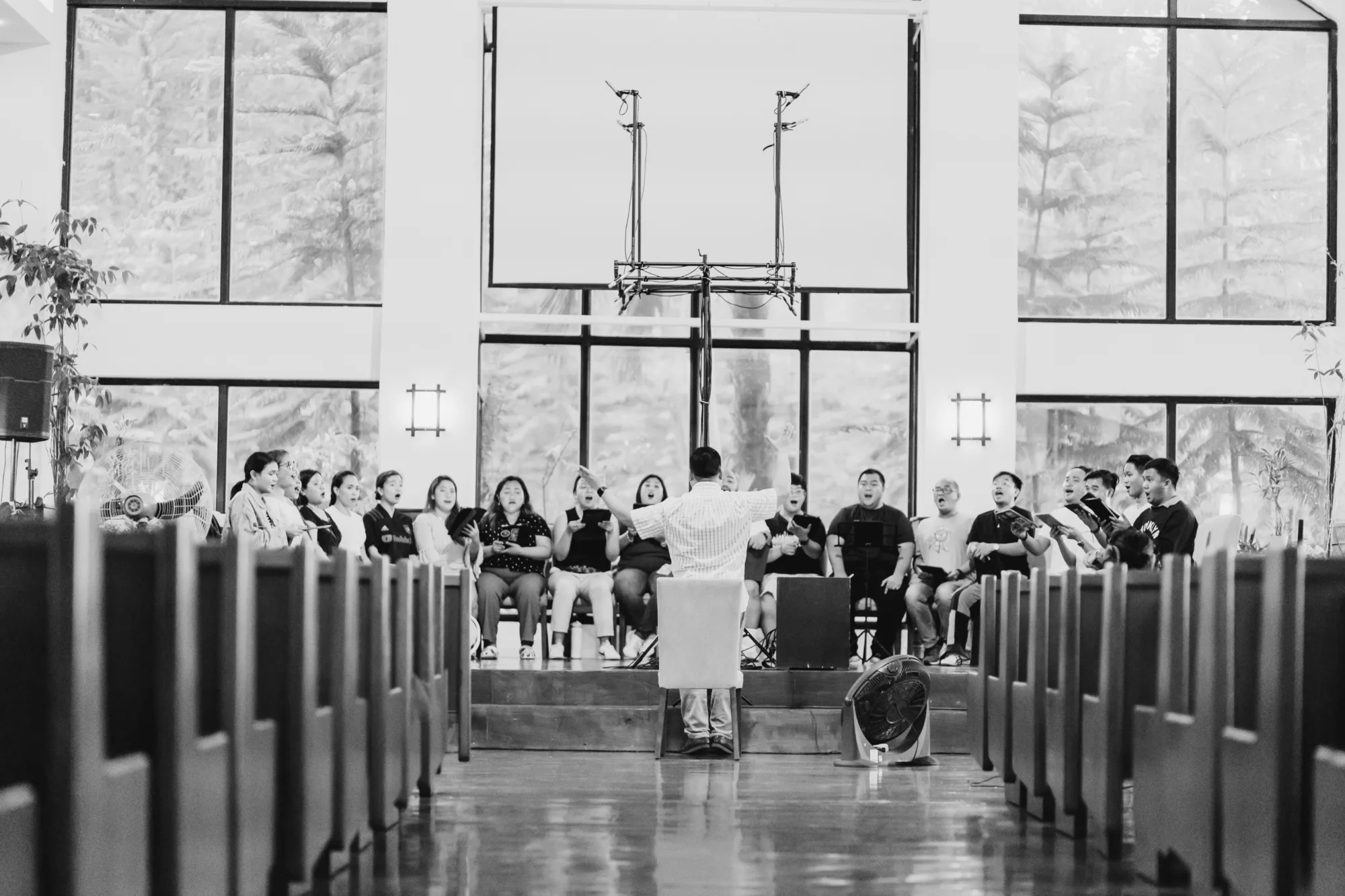 A conductor leads a choir in a bright church hall, seen from behind the pews